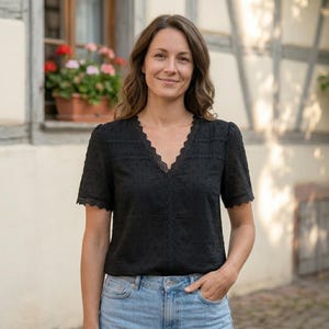Op de afbeelding: Een zwarte blouse met korte mouwen, een V-hals en kanten details. De blouse heeft een subtiele stippenstructuur en wordt gecombineerd met lichtblauwe jeans. De vrouw lacht, met haar hand in haar zak.