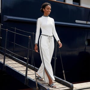 Puede incluir: Una camiseta blanca de manga larga con cuello alto y pantalones anchos a juego con detalles de rayas negras y blancas en los laterales. Los pantalones incluyen una cintura con cordón y aberturas laterales en las piernas. Se combina con sandalias.