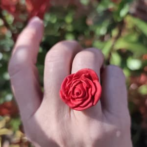 Puede incluir: Un anillo de rosa roja, hecho de un material similar a la arcilla, se muestra en una mano. El anillo presenta una rosa detallada con intrincadas formaciones de pétalos. El fondo tiene follaje verde borroso y hojas rojas.