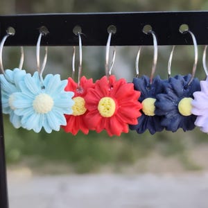 Puede incluir: Pendientes de aro con diseño de flores de margarita en varios colores, incluyendo azul claro, rojo y azul marino. Cada pendiente presenta un aro plateado y una margarita con centro amarillo. Los pendientes se exhiben en un soporte negro.