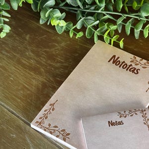 May include: A stack of light pink notepads with the word "Nototas" printed in brown. A smaller notepad with the word "Notas" is also visible. Each notepad has a brown floral corner design. Greenery is in the background.