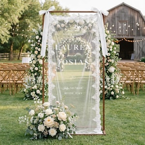 May include: A rectangular wooden frame holding a sheer white fabric panel with lace trim and white ribbons at the top corners. The text reads: WELCOME TO OUR FOREVER, LAUREN and JOSIAS, July 01 2026, BUCINE, ITALY. A floral arrangement sits at the base.