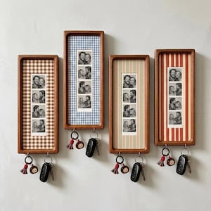May include: Four rectangular wooden wall-mounted key holders with patterned backings. Each frame displays a vertical strip of four black and white photos. Two metal hooks at the bottom of each frame hold key rings with tassels and charms.