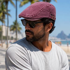 May include: A burgundy newsboy cap with a curved brim and a small, adjustable strap. The cap is worn with black sunglasses and a gray t-shirt. The background features palm trees and a beach scene.