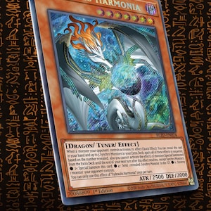 Op de afbeelding: Een ruilkaart getiteld Fydraulis Harmonia, met een witte draak met oranje haar en een gloeiende bol. De kaart is een Dragon Tuner Effect-monster met 2500 ATK en 2000 DEF, gemarkeerd als 1st Edition, BLZD-EN024, inclusief gedetailleerde speltekst.