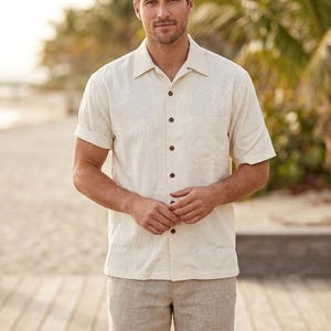Puede incluir: Una camisa crema de manga corta con un sutil estampado de palmeras, combinada con pantalones cortos de color beige claro. La camisa tiene una botonadura frontal y un bolsillo en el pecho. El atuendo es adecuado para climas cálidos.