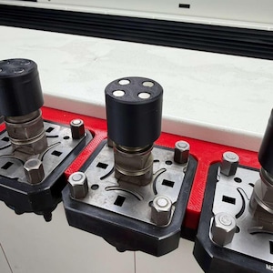 May include: Close-up of industrial machinery components. The image shows several black cylindrical parts with four white dots on top, mounted on metal plates with bolts and nuts. The plates are set on a red base, with a white and black background.
