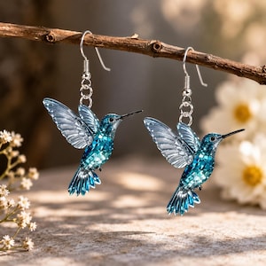 Peut inclure: Une paire de boucles d'oreilles pendantes avec des breloques colibri bleues à facettes. Chaque oiseau possède des ailes translucides et un corps texturé et scintillant dans des tons bleu sarcelle et azur. Les breloques pendent à des crochets en métal argenté.