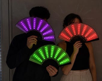 Puede incluir: Tres abanicos plegables de mano con marcos negros y luces LED integradas. Un abanico brilla en color morado, otro en rojo y otro en verde. Los abanicos están fabricados con paneles de tela translúcida que se iluminan al encender las luces.