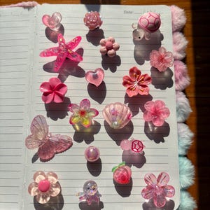 Puede incluir: Una colección de pequeños dijes decorativos rosas con diversas formas, incluyendo flores, corazones, una mariposa, una estrella de mar, una concha, una tortuga, un pulpo y un melocotón, dispuestos sobre las páginas rayadas de un cuaderno abierto.