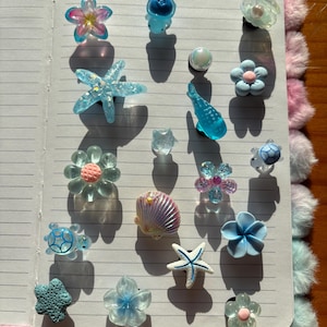 Puede incluir: Una colección de dijes decorativos con diversas formas, incluyendo flores, criaturas marinas como tortugas y estrellas de mar, y conchas. Los dijes están realizados en tonos azules, rosas y blancos con acabados translúcidos, nacarados y texturizados.