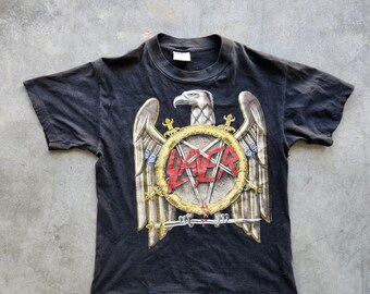 May include: Black t-shirt featuring a large graphic of an eagle with outstretched wings, a circular emblem, and the word "SLAYER" in red. The design has a vintage, distressed look. The shirt is short-sleeved and has a crew neck.