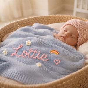 May include: A sleeping baby wearing a pink knit hat rests in a woven basket. The baby is covered with a light blue blanket embroidered with the name "Lottie", flowers, a rainbow, and a heart.