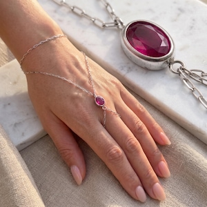 Op de afbeelding: Een zilveren handketting met een kleine, ovaal geslepen donkerroze edelsteen, verbonden door een fijne ketting aan een polsband. Een bijpassende zilveren ketting met een grote, ovaal geslepen donkerroze hanger ligt op een witte marmeren ondergrond.