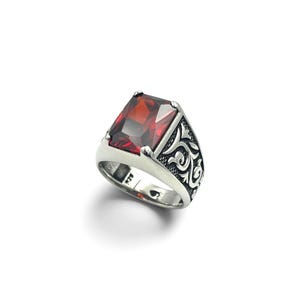 May include: A silver ring featuring a large, rectangular, faceted deep red garnet-colored stone set in a prong setting. The band is crafted from silver with intricate, raised scrollwork patterns along the sides and a 925 hallmark stamp inside.