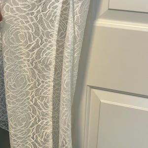 May include: A white lace fabric with a floral rose pattern. The delicate material is draped, revealing the intricate design and texture. The fabric is likely used for clothing or home decor.