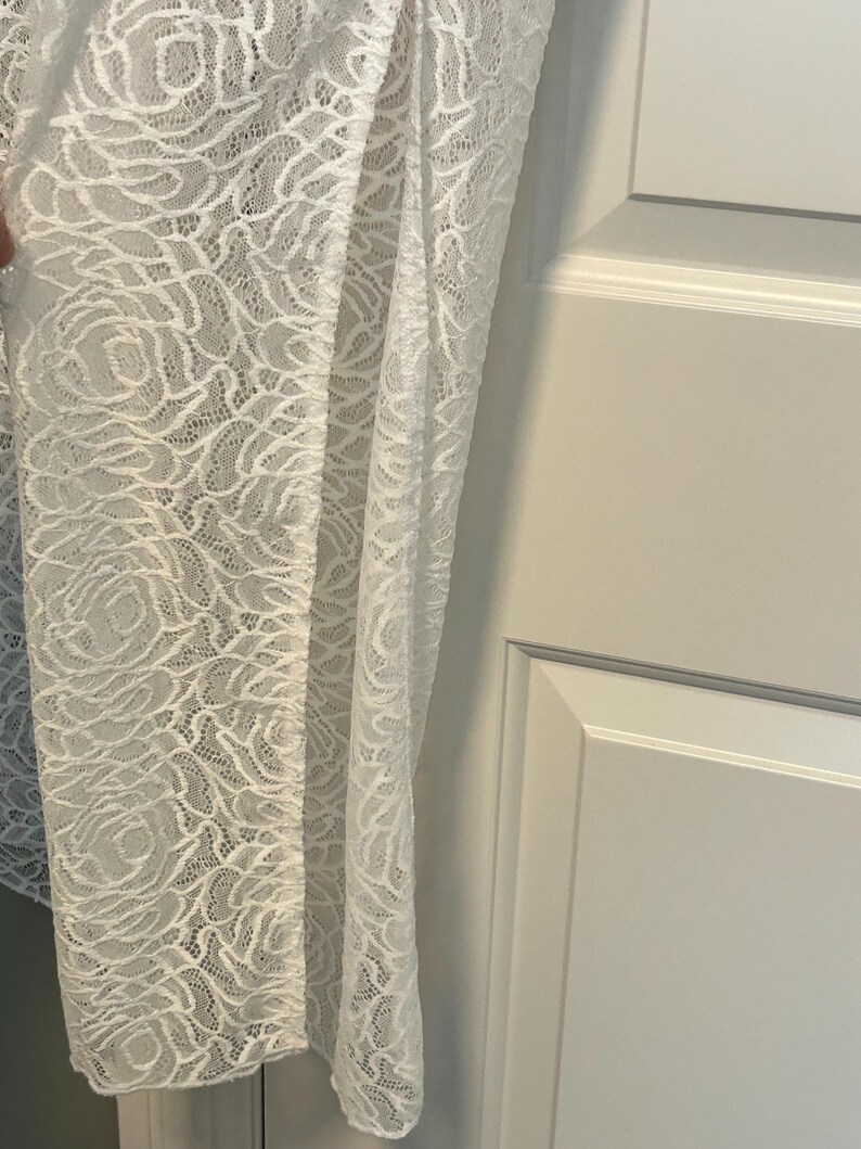 May include: A white lace fabric with a floral rose pattern. The delicate material is draped, revealing the intricate design and texture. The fabric is likely used for clothing or home decor.