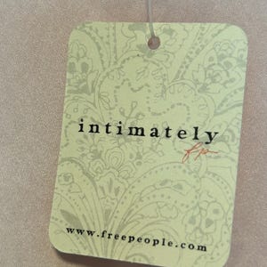 May include: A rectangular tag with rounded corners, featuring the text "intimately" in a stylized font and the initials "fp" in red. The tag has a light green background with a floral pattern and the website address www.freepeople.com.