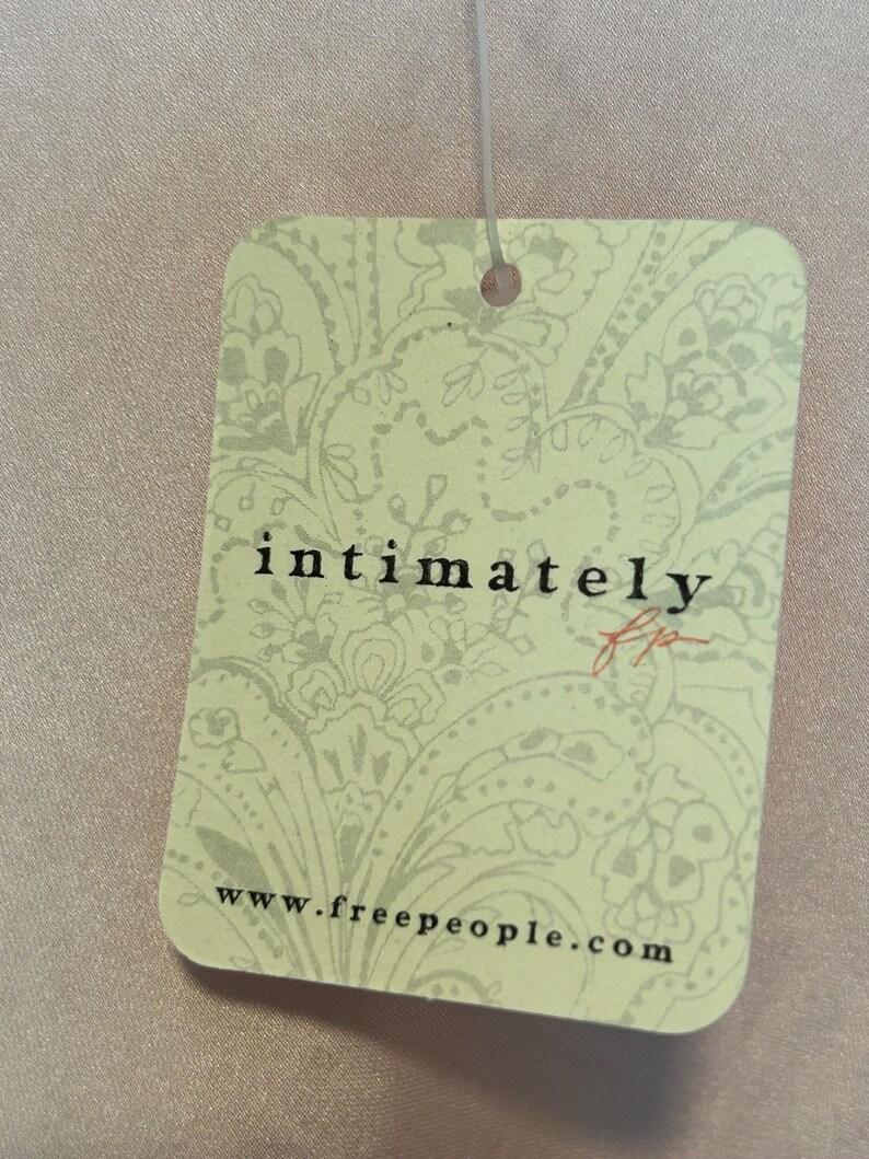 May include: A rectangular tag with rounded corners, featuring the text "intimately" in a stylized font and the initials "fp" in red. The tag has a light green background with a floral pattern and the website address www.freepeople.com.