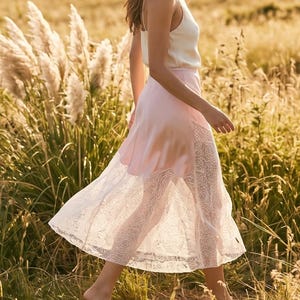 May include: A woman wearing a white camisole top and a light pink skirt with a white lace hem, walking through a field of tall grass. The skirt flows in the wind, creating a soft, feminine silhouette.