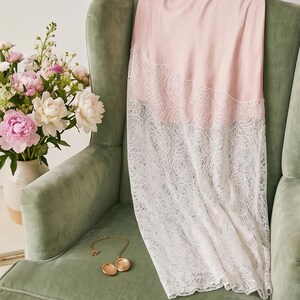 May include: A pale pink slip dress with a white lace overlay, draped over a green armchair. A gold locket necklace rests on the chair. A vase of pink and white peonies sits nearby, creating a soft, feminine aesthetic.