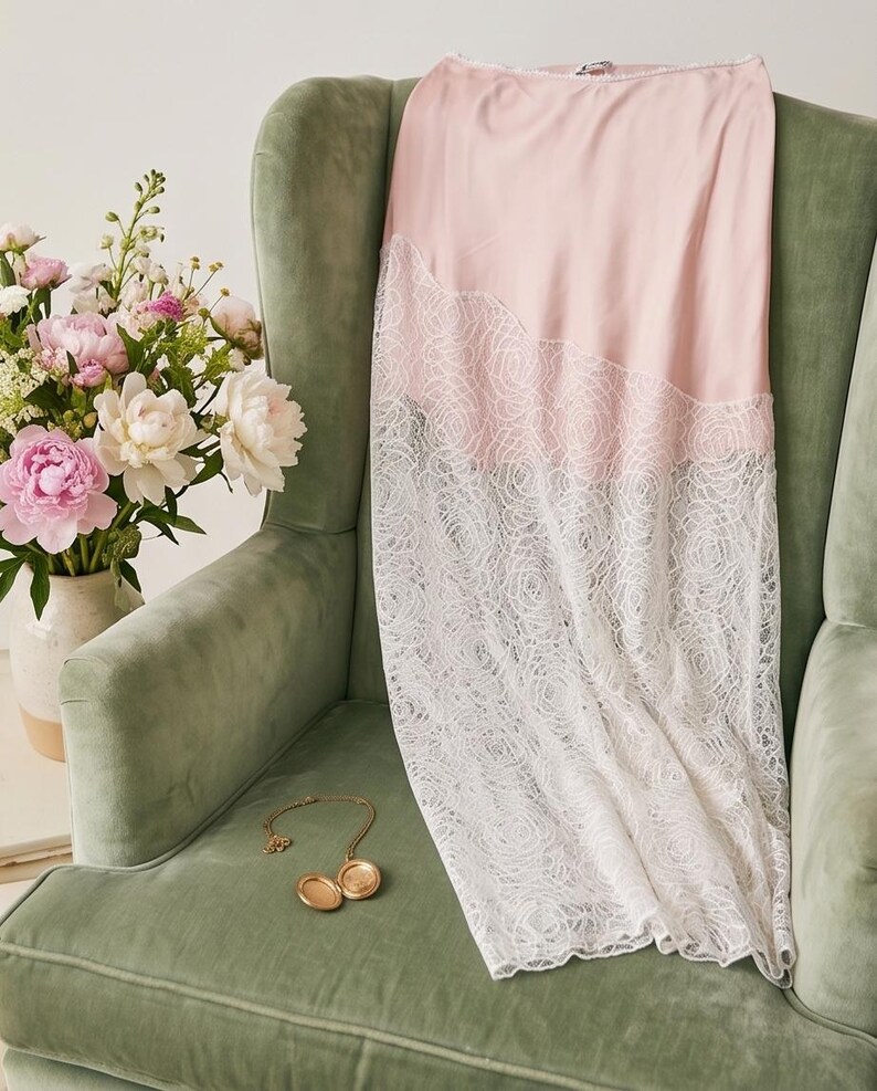 May include: A pale pink slip dress with a white lace overlay, draped over a green armchair. A gold locket necklace rests on the chair. A vase of pink and white peonies sits nearby, creating a soft, feminine aesthetic.