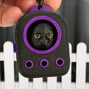 May include: A black and purple 3D-printed keychain featuring a rounded rectangular housing with a circular window. Inside the window is a sculpted black cat head with prominent yellow eyes. The device includes three small purple circular accents.