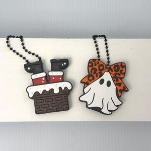May include: Two silicone charms with black ball chains. One charm depicts Santa's legs in red pants and black boots emerging from a brown brick chimney with white snow. The other charm is a white ghost wearing an orange bow with black leopard spots.