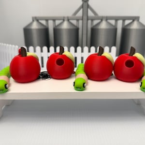 May include: Four red apple-shaped objects with brown stems and small holes on the sides. Each apple is paired with a green segmented worm figure that has a light pink face and black eyes, creating a set of decorative items with a smooth finish.