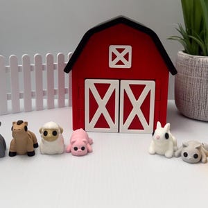 May include: A collection of small animal-shaped erasers including a chicken, cow, goat, horse, sheep, pig, rabbit, cat, chick, highland cow, and squirrel, arranged in front of a red and white wooden barn model and a white picket fence.