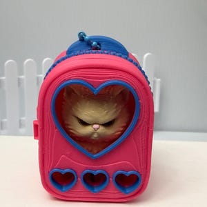 May include: A pink cat carrier with a blue top and heart-shaped cutouts. The carrier features a heart-shaped window displaying a fluffy cat. Three heart-shaped cutouts are at the bottom. The cat has an angry expression.