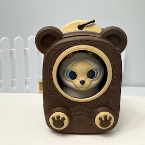 May include: A brown, bear-shaped container with a textured surface and two small paw prints at the base. Inside the circular opening sits a small, cream-coloured creature with large, bright blue eyes, a dark nose, and small ears.
