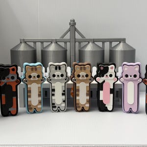 May include: A collection of eight small, stylized cat-shaped figurines standing in a row. Each cat features a unique color pattern, including gray, orange, brown, black, white, and lavender, with simple facial features and small paws.