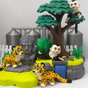 May include: A jungle-themed playset featuring two yellow and black spotted leopard figures, two white and black monkey figures, a gray stone-style totem statue, a brown tree with green leaves, a blue waterfall feature, and a green grassy base.