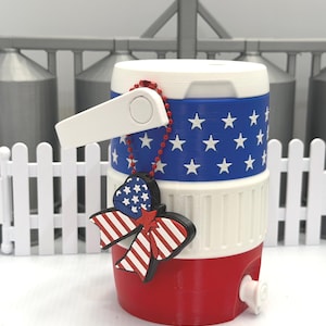May include: A small cylindrical beverage cooler featuring a red, white, and blue color scheme with a white star pattern on a blue band. A red beaded chain attaches a bow-shaped charm with a star and stripe pattern to the white handle.