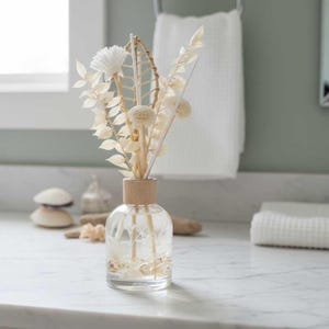 May include: A clear glass diffuser bottle with a wooden collar, filled with seashells and reeds. The bottle holds dried flowers and decorative elements. The arrangement sits on a white marble countertop, suggesting a spa or bathroom setting.