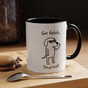 May include: White ceramic mug with a black interior and handle. The mug features a cartoon dog wearing sunglasses and giving the middle finger, with the text "Go fetch... Yourself". A spoon and a macaron are in the foreground.