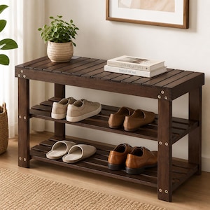 May include: A dark brown wooden shoe rack bench with three slatted tiers. The top surface holds a potted green plant and two stacked books titled Live Beautiful and This Is Home. The shelves contain four pairs of shoes including sneakers and loafers.