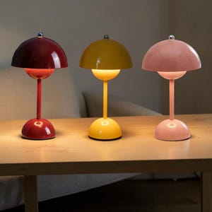 Pode incluir: Três candeeiros de mesa coloridos com design retro. Cada candeeiro tem um abajur e base arredondados, com um caule fino. Os candeeiros são vermelhos, amarelos e rosa, colocados numa mesa de madeira.