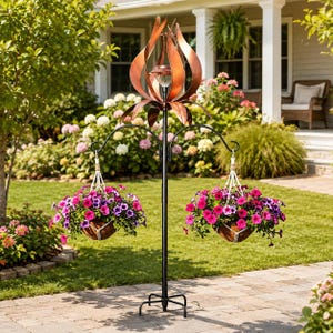 May include: A black metal garden plant stand featuring a central pole with a copper-colored kinetic wind spinner at the top and two curved arms holding hanging baskets filled with vibrant pink and purple petunia flowers on white ropes.
