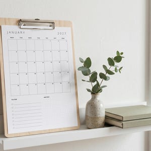 May include: A light wood clipboard holding a white January 2027 calendar page with a grid layout, a to-do list, and a notes section. Next to it sits a small speckled ceramic vase containing green eucalyptus stems and two stacked sage green books.