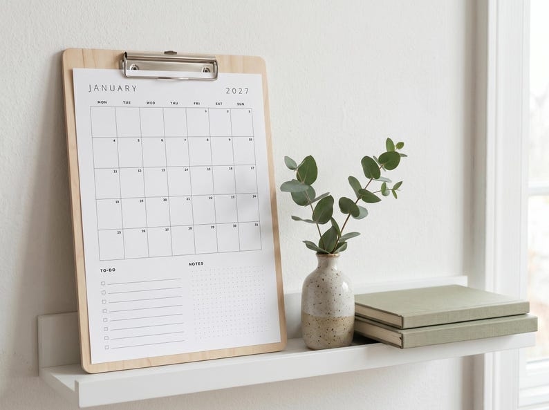 May include: A light wood clipboard holding a white January 2027 calendar page with a grid layout, a to-do list, and a notes section. Next to it sits a small speckled ceramic vase containing green eucalyptus stems and two stacked sage green books.