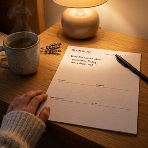 May include: A notepad with the words "Brain Dump" and a list of worries is on a wooden nightstand. A pen rests on the notepad. A mug of tea and a lamp are also on the nightstand.