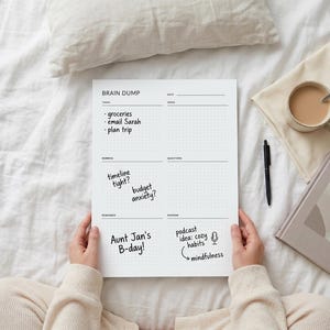 May include: A white Brain Dump notepad with dot grid paper is held by a person. The notepad has sections for tasks, worries, questions, and more. Handwritten notes include "groceries," "email Sarah," and "plan trip." A pen, a book, and a cup of coffee are also visible.