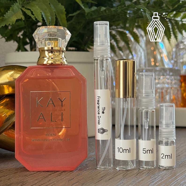 May include: A coral-colored perfume bottle with a gold cap and a clear, faceted top. The bottle is labeled "KAYALI EDEN SPARKLING LYCHEE 19". Beside it are several clear glass spray bottles labeled "The Fragrance Drop", and "10ml", "5ml", and "2ml".