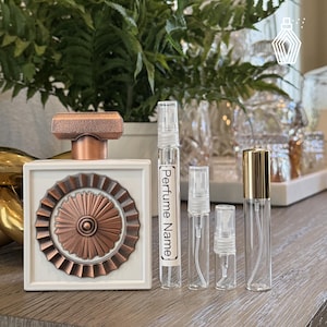 May include: A white and copper perfume bottle with a decorative design, next to several clear glass perfume sample vials. One vial has the text "Perfume Name". The bottles are arranged on a wooden surface.