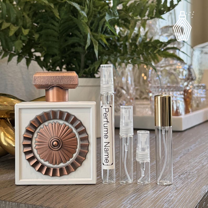 May include: A white and copper perfume bottle with a decorative design, next to several clear glass perfume sample vials. One vial has the text "Perfume Name". The bottles are arranged on a wooden surface.