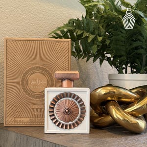 May include: A white perfume bottle with a copper-colored decorative element and a matching cap. The bottle is next to a box with Arabic text and a sunburst design. The bottle contains 100 ml of perfume.