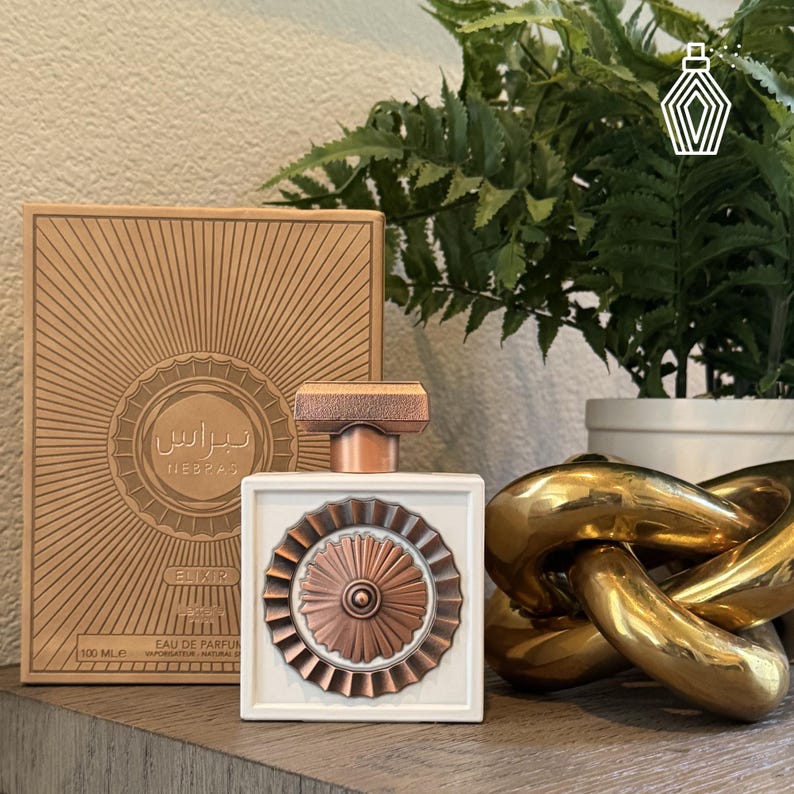May include: A white perfume bottle with a copper-colored decorative element and a matching cap. The bottle is next to a box with Arabic text and a sunburst design. The bottle contains 100 ml of perfume.