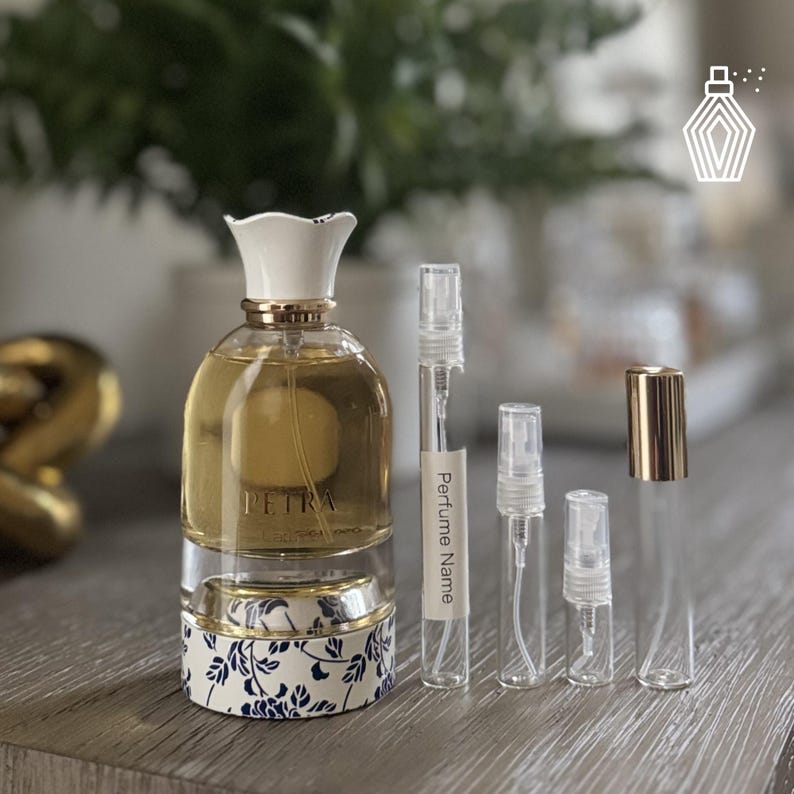 May include: A perfume bottle with a white floral-shaped cap and gold detailing, filled with a light amber liquid. The bottle sits on a base with a blue and white floral design. Several small, clear sample vials are next to the bottle.